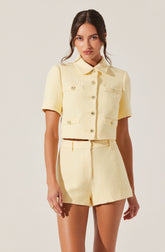 A woman is wearing a light yellow, textured, short-sleeved cropped jacket with a collar and decorative buttons on the front and pockets. She is also wearing matching high-waisted shorts made of the same fabric.